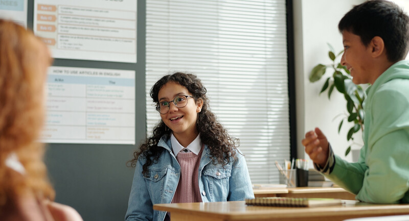 A group of multilingual learners sit in a classroom, talking and smiling together.