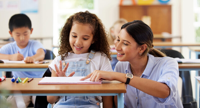 A teacher and student talking to each other at a desk while looking closely at a notebook together